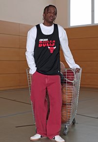 Fekete kosárlabda mez a "Chicago Bulls" logójával, piros széles szárú nadrággal és fehér tornacipővel párosítva; egy kosárlabdákat tartalmazó drótkocsi mellett áll.