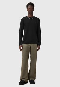 Schwarz langärmliges Shirt mit subtilen Logo-Detail, kombiniert mit lockeren olivgrünen Hosen und schwarzen strukturierten Schuhen.