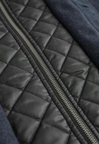 Geprägter schwarzer Stoff mit einer glatten Textur, der in der Mitte einen silbernen Reißverschluss aufweist, der auf einer Seite von einem strukturierten blauen Material gesäumt wird.