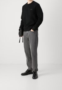 Osoba ubrana w czarną bluzę z logo "BOSS", szare dżinsy, czarne skórzane buty, trzymająca małą czarną torbę na ramię, stojąca na jednolitym białym tle.