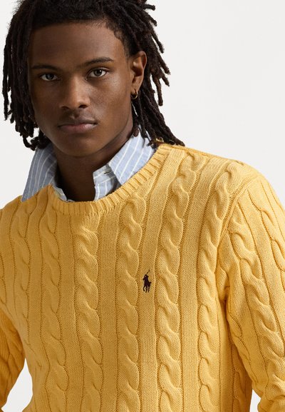 Un pull en maille côtelée jaune avec un col rond, présentant un petit logo sur la poitrine et associé à une chemise rayée bleu et blanche.