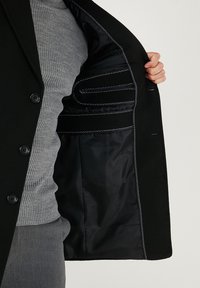 Czarny płaszcz z gładką podszewką; posiada zapięcie na guziki, szary prążkowany sweter pod spodem oraz kontrastujące przeszycia na podszewce.