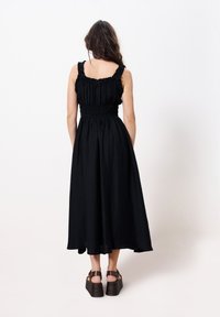 Schwarzes Midi-Kleid aus strukturiertem Stoff, mit Rüscheträgern und smockiertem Taillenbereich. Kombiniert mit braunen Plateausandalen. Rückansicht.