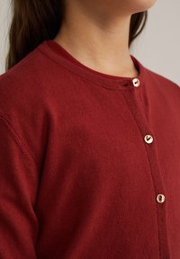 Läheltä kuvattu henkilö, jolla on päällään punainen neulevillatakki kullanvärisillä nappeilla, näkyy ylärinta ja kaula-alue.