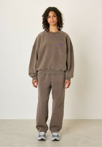 Prevelik rjav pulover in usklajeni trenirke z vijoličastim logotipom. Mehak material s rebrastimi manšeti in pasom. Vključeni so športni čevlji.