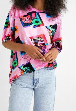 Person iført lyseblå jeans og en løs lyserød grafisk skjorte med farverige abstrakte former og delvist synlig tekst.