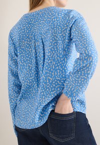 Femme portant une chemise à manches longues ample à motif bleu, avec la main dans la poche arrière d'un jean en denim foncé, tournée de dos.