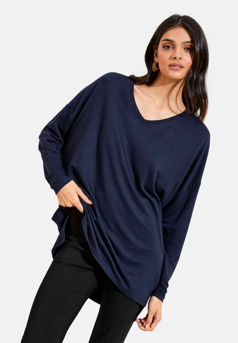 Majica tamnoplave boje s dugim rukavima i V-izrezom, izrađena od mekanog materijala, s opuštenim krojem i zakrivljenim rubom, u kombinaciji s crnim hlačama.