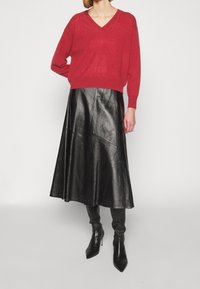 Kvinna iförd en röd V-ringad tröja, svart läder midikjol och svarta klackboots som går upp till knäna, stående mot en enfärgad vit bakgrund.