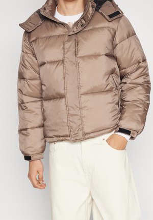 Mężczyzna ubrany w puchową kurtkę koloru taupe z czarnymi akcentami na białym swetrze, w połączeniu z kremowymi spodniami, ręka w kieszeni, na jednolitym tle.