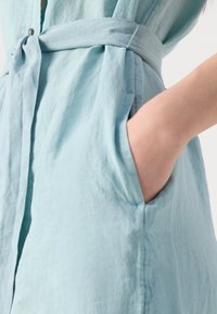 Šviesiai mėlyna lininė suknelė su surišamu juosmeniu, turi šonines kišenes ir mygtukų užsegimą. Medžiaga turi švelnų, tekstūruotą išvaizdą.