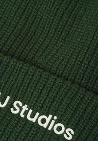 Zielona dziana czapka beanie o prążkowanej fakturze. Z przodu znajduje się wyraźnie widoczny biały nadruk z logo "MJ Studios".