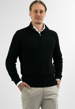 Mann trägt schwarzen Pullover mit kurzem Reißverschluss über weißem Hemd und beiger Hose, steht mit Händen in den Taschen vor einfarbigem Hintergrund.