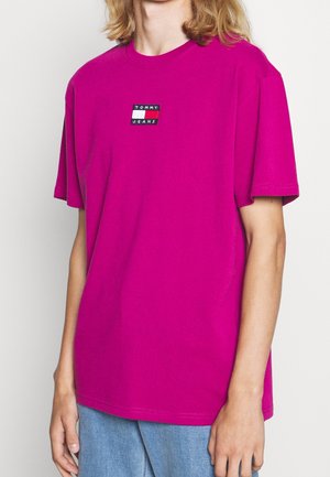 Osoba ubrana w jasnoróżowy t-shirt z krótkim rękawem z małym logo Tommy Jeans na piersi, w połączeniu z jasnoniebieskimi dżinsami.