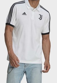 Homme portant un polo blanc Adidas avec le logo noir de la Juventus et des bandes sur les manches, associé à un jean bleu clair et un accessoire en chaîne.