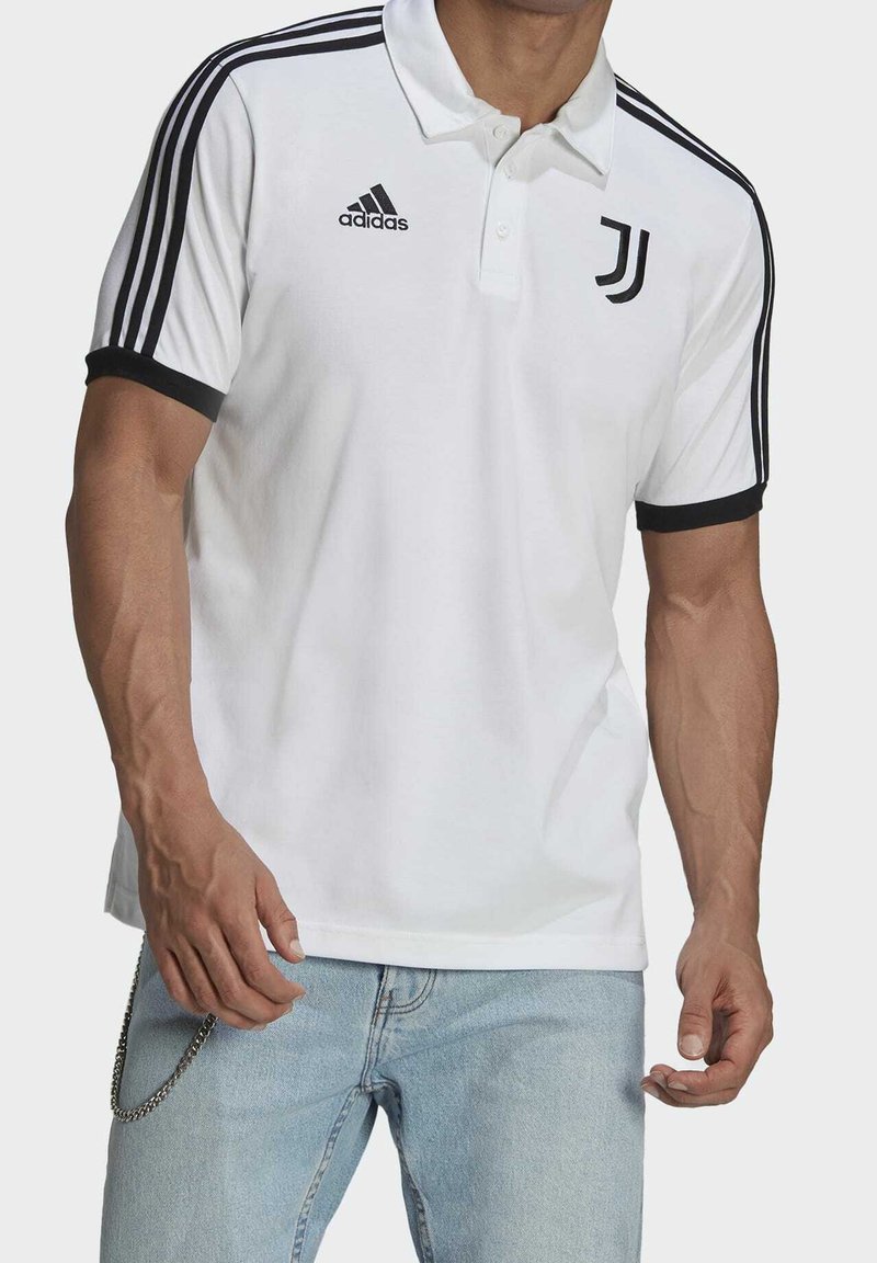 Homme portant un polo blanc Adidas avec le logo noir de la Juventus et des bandes sur les manches, associé à un jean bleu clair et un accessoire en chaîne.