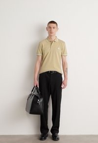 Khaki polo krekls ar baltu apkakli, melnas bikses un melnu rokassomu. Modelis stāv uz vienkārša baltā fona.
