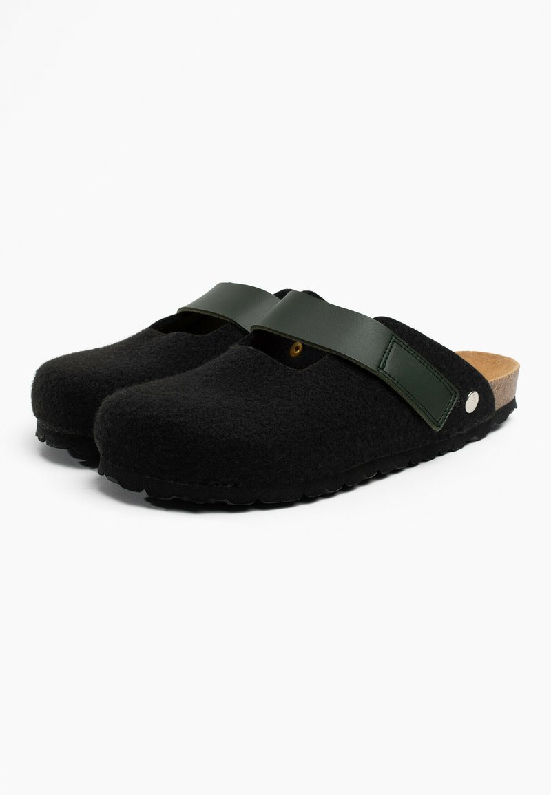 Zuecos negros tipo slip-on con parte superior texturizada, correa ajustable en verde oscuro y plantilla de corcho, presentados sobre un fondo blanco.