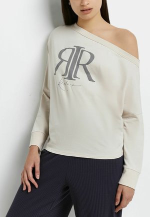 Kobieta ubrana w kremową bluzę z odkrytymi ramionami z napisem "RIR Relax" oraz ciemne prążkowane spodnie, stojąca na tle białej ściany.