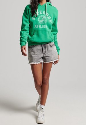 Persoon draagt een groene hoodie met de tekst "Superdry ATH.DEN", grijze denim shorts met rafels, witte high-top sneakers, lopend naar voren.