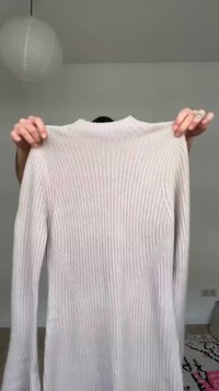 Persona che tiene in mano un maglione beige a maniche lunghe, lavorato a coste, davanti a una parete semplice con una lanterna di carta appesa sullo sfondo.