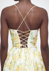 Πίσω όψη ατόμου που φοράει κίτρινο φόρεμα με λουλούδια, με λεπτούς χιαστί ιμάντες και ραφές που δένονται στην ανοιχτή πλάτη.