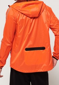 Personne portant une veste imperméable orange vif avec capuche et poche zippée noire dans le bas du dos, associée à un pantalon noir.