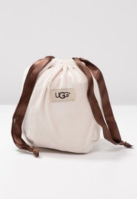 Miękka, kremowa, sznurowana torba z korduroyu, z brązowymi satynowymi wstążkami i naszywką z logo UGG z przodu, na jednolitym tle.