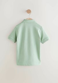 Polo-shirt vert clair à manches courtes avec un col côtelé et un tissu texturé, présenté sur un cintre en bois contre un fond clair.