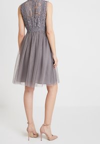Robe en tulle gris avec corsage en dentelle, design sans manches et jupe mi-longue. Le mannequin porte des talons beiges à bride cheville, dévoilant les jambes.