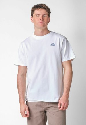 Jonge man die een wit T-shirt draagt met de tekst "Good Morning Sunshine" op de borst, gecombineerd met lichtbruine broek, staand tegen een effen achtergrond.