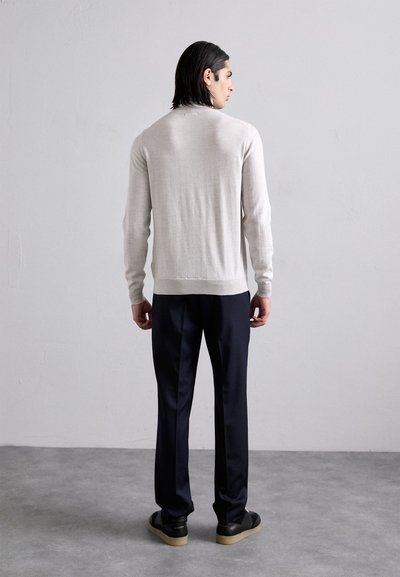 Maglione a maglia di un beige chiaro con maniche lunghe, abbinato a pantaloni blu scuro e scarpe nere. Texture liscia e design semplice.