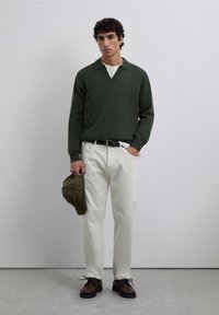 Jersey verde con cuello, camiseta blanca, pantalones beige, zapatos marrones y una gorra de cuadros en la mano. Texturas suaves en todo.