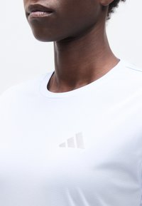 Κοντινό πλάνο ενός ατόμου που φοράει λευκό μπλουζάκι Adidas με διακριτικό λογότυπο στο στήθος, δείχνοντας τον λαιμό και το κάτω μέρος του προσώπου.