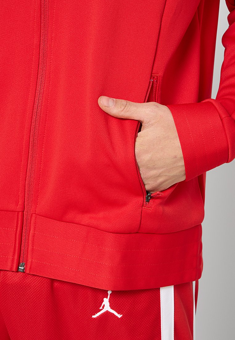Chaqueta atlética roja con un bolsillo con cremallera, dobladillo acanalado y detalle del logo en blanco en la manga. Fabricada con una tela suave y texturizada.