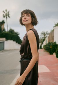 Jeune femme aux cheveux bruns courts portant une robe sombre sans manches, debout à l'extérieur sur un chemin proche de plantes vertes et de palmiers.
