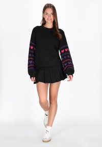 Czarny dzianinowy sweter z kolorowymi wzorami serc na rękawach, w zestawieniu z czarną plisowaną spódnicą i białymi trampkami. Całość ma gładką teksturę.