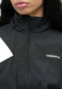 Svart glidelåsjakke laget av lett materiale. Har høy krage og brodert hvitt Adidas-logo på venstre bryst.