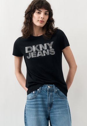 Młoda kobieta z kręconymi włosami, ubrana w czarny t-shirt "DKNY JEANS" i niebieskie jeansy, stoi z rękami w kieszeniach na jednolitym tle.