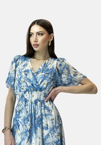 Robe florale bleue avec des manches transparentes bouffantes, taille froncée et accents dorés. Présente un motif avec une texture légère et aérée.