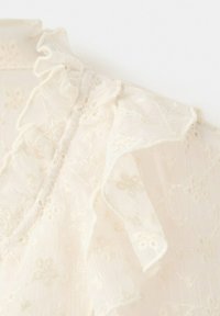 Tissu en dentelle blanche transparente avec broderie florale et bordure volantée sur un col haut, mettant en valeur une texture délicate et une couture complexe.