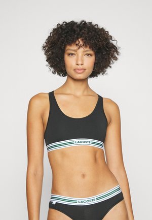 Schwarzer Sport-BH mit einem breiten Bund, der ein strukturiertes Lacoste-Logo und grüne Streifen aufweist. Der Stoff ist dehnbar und glatt für maximalen Komfort.