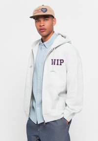 Gray zip-up hoodie with "WIP" embroidered in purple, worn over a light blue textured button-up shirt, paired with a tan logo cap.