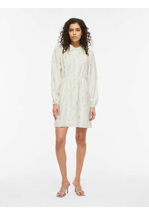 Nő áll fehér, hosszú ujjú, virágmintás ruhában és ezüst színű, magas sarkú szandálban, egyszínű fehér háttér előtt.