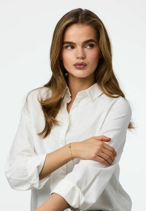 Chemise blanche à boutons avec manches retroussées, main du modèle reposant sur son bras, portant un délicat bracelet en or avec une petite pierre précieuse.