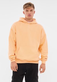 Felpa con cappuccio color pesca realizzata in morbido cotone misto, presenta una tasca frontale, maniche lunghe e una vestibilità comoda con polsini e bordo a coste.