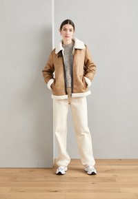 Giacca in suede marrone con colletto e polsini in shearling bianco, chiusura con zip, indossata sopra un maglione grigio a trama; abbinata a pantaloni bianco sporco e sneakers.