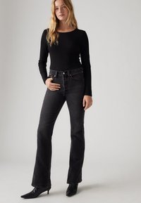 Haut noir à manches longues avec un décolleté arrondi associé à un jean évasé taille haute en délavé foncé. Mannequin portant des bottes à talons aiguilles noires.