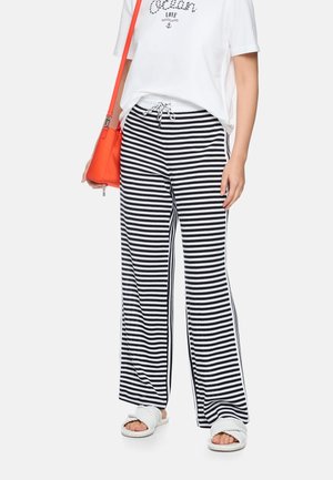 Frau trägt weißes kurzes T-Shirt mit Text, schwarz-weiß gestreifte weite Hose mit weitem Bein, weiße Pantoletten und trägt eine orangefarbene Schultertasche.