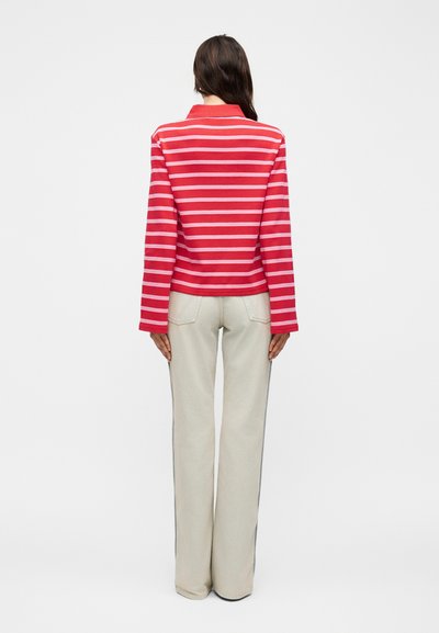 Γυναίκα που στέκεται με την πλάτη, φοράει κόκκινο και λευκό ριγέ μακρυμάνικο πουκάμισο και ανοιχτό μπεζ παντελόνι με φαρδιά πόδια σε απλό φόντο.